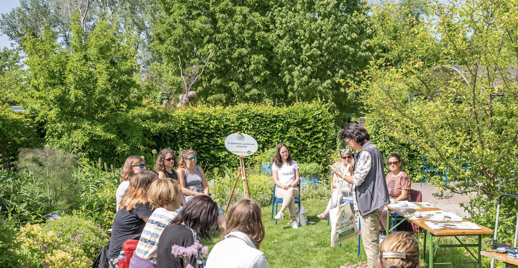 Veranstaltungsfoto für Fachtagung Gartenpädagogik 30.05.26 in Tulln. In einem Garten mit Hecken und Bäumen sitzen mehrere Frauen und reden miteinander. Rechts stehen zwei Holztische mit Materalien