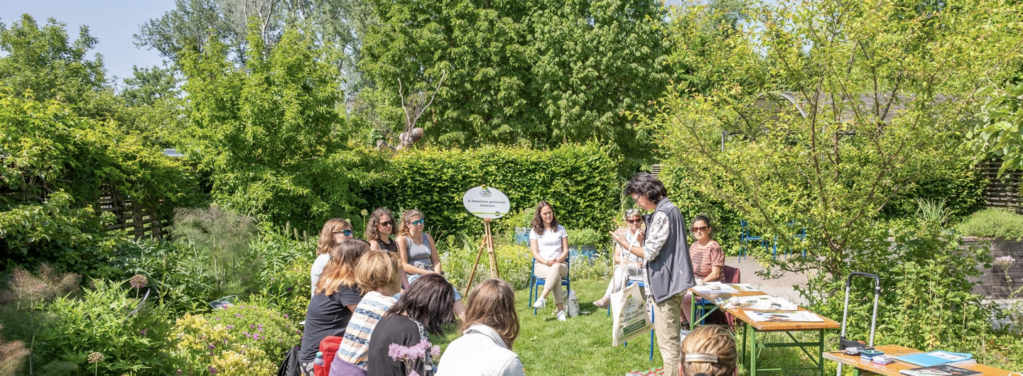 Veranstaltungsfoto für Fachtagung Gartenpädagogik 30.05.26 in Tulln. In einem Garten mit Hecken und Bäumen sitzen mehrere Frauen und reden miteinander. Rechts stehen zwei Holztische mit Materalien